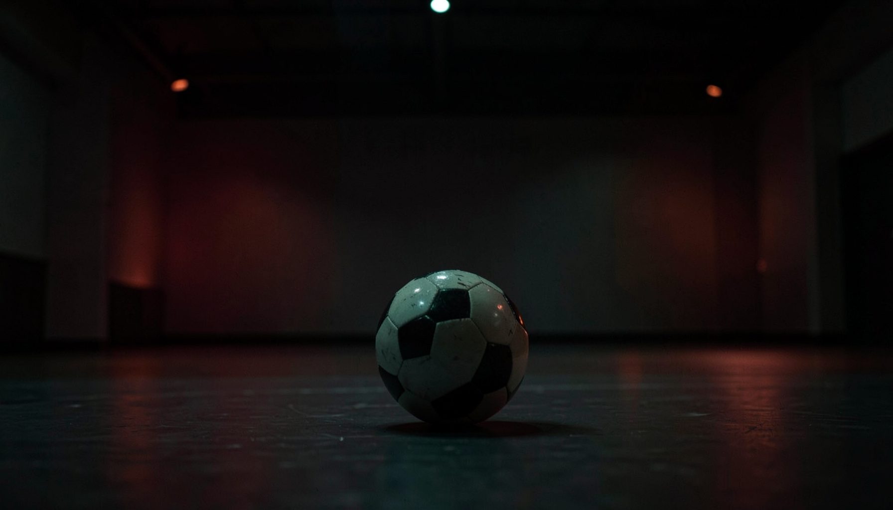 Tamamen karanlık bir spor salonunda, loş yeşil ve turuncu ışık huzmeleri altında bir futbol topunun zeminde yuvarlanışı